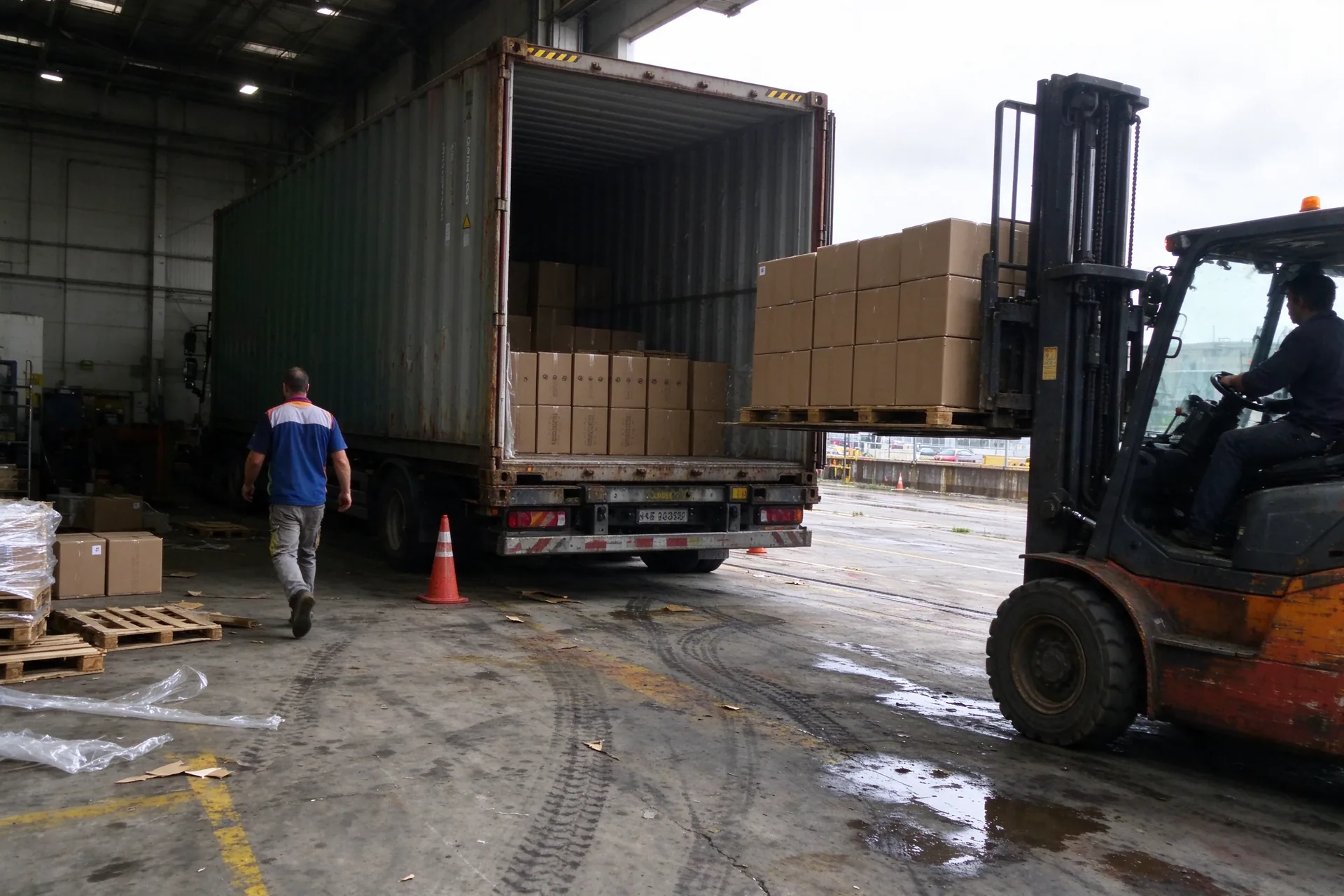 Entrepôt près d'Anvers : un chariot élévateur sort une palette de cartons d'un conteneur à quai, sol mouillé, temps gris.